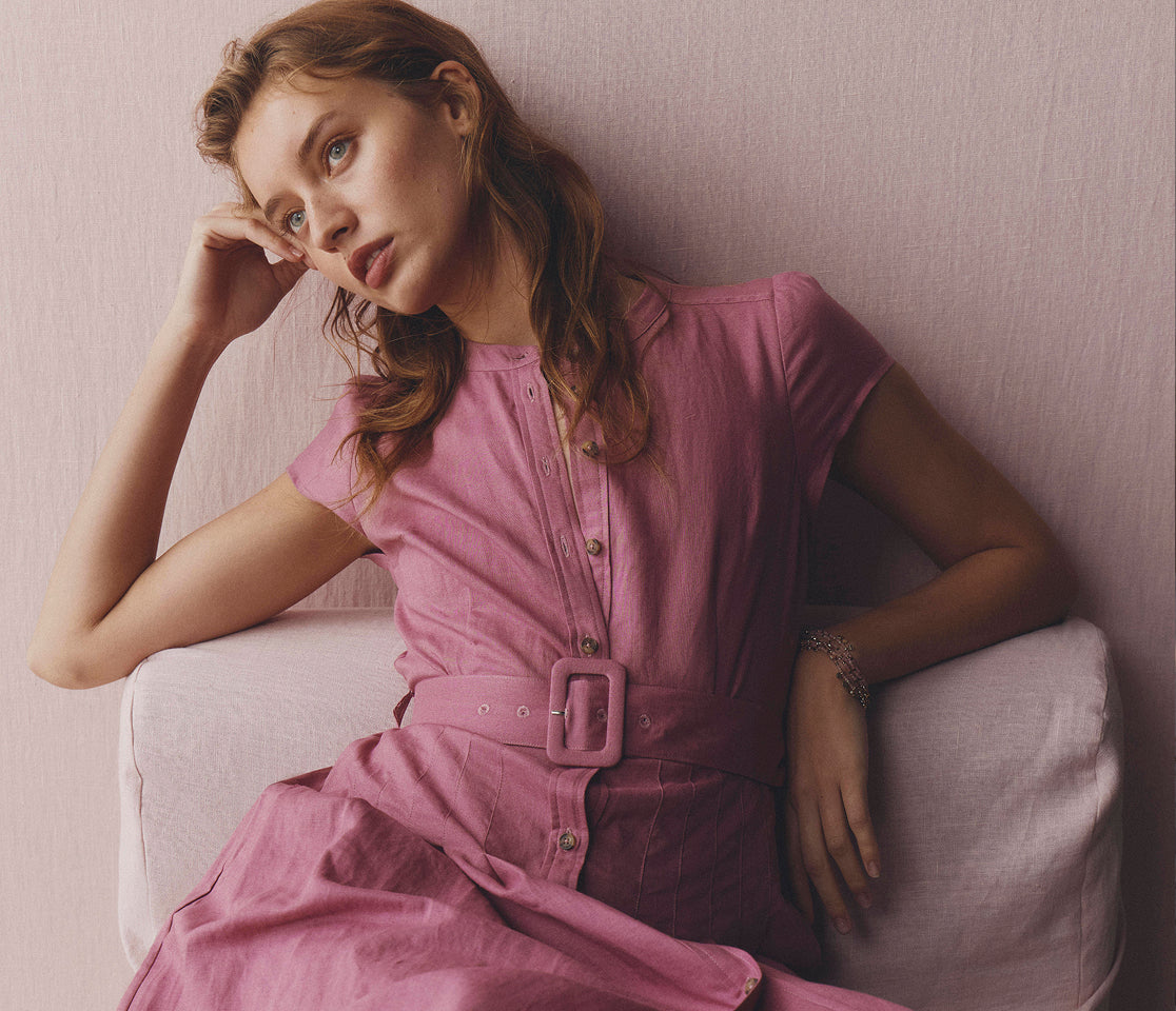 Woman in a pink dress sitting on a white couch against a light pink wall.