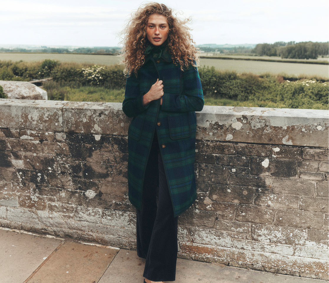 Person with long, curly hair wearing a dark green and navy tartan coat and wide-leg trousers, standing against a weathered stone wall with open countryside and hedgerows in the background. The outdoor fashion portrait is set in a rural landscape under soft, overcast light.