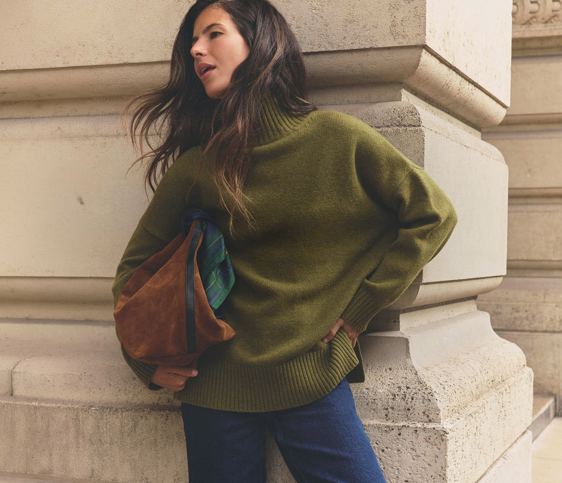 Person leaning against a stone building, wearing an oversized olive-green turtleneck sweater and dark blue jeans, while carrying a brown suede tote bag with a green tartan scarf tucked inside. The outfit is set against a neutral architectural backdrop, creating a relaxed, heritage-inspired look.