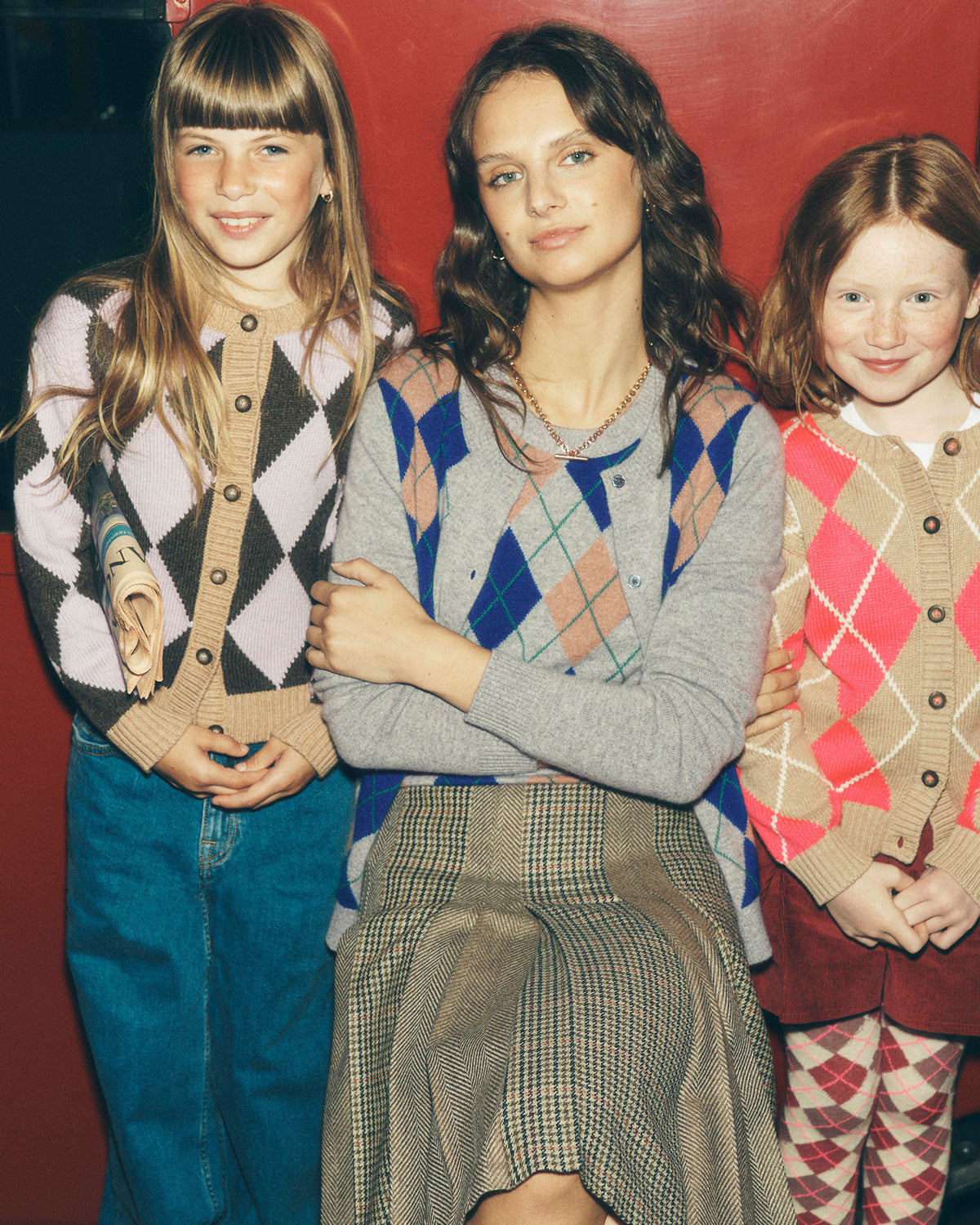 Three women posing together in vintage argyle cardigans and skirts against a red background.