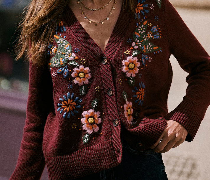 A close-up of a woman wearing a burgundy cardigan sweater with colorful floral embroidery on the front, paired with dark jeans and layered gold necklaces.