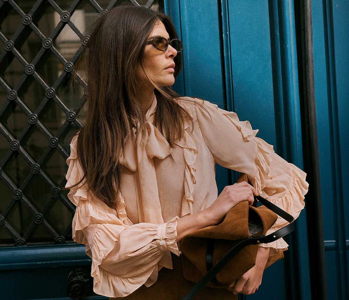 A woman with long dark brown hair wearing a beige ruffled blouse and sunglasses, holding a brown leather handbag against a blue door background.
