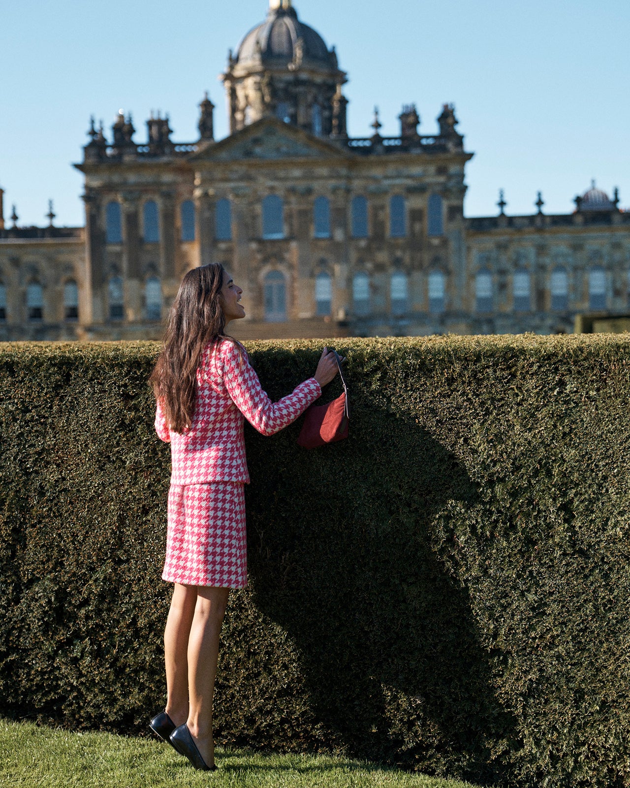 A woman in a pink and white checkered suit stands in front of a tall hedge, holding a red handbag, with a grand historic building in the background under a blue sky.