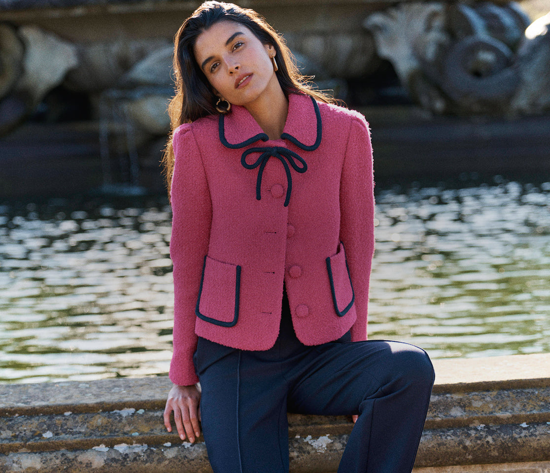 A woman sits on a stone ledge in front of a fountain, wearing a pink textured jacket with black trim and a bow at the collar, paired with dark trousers.