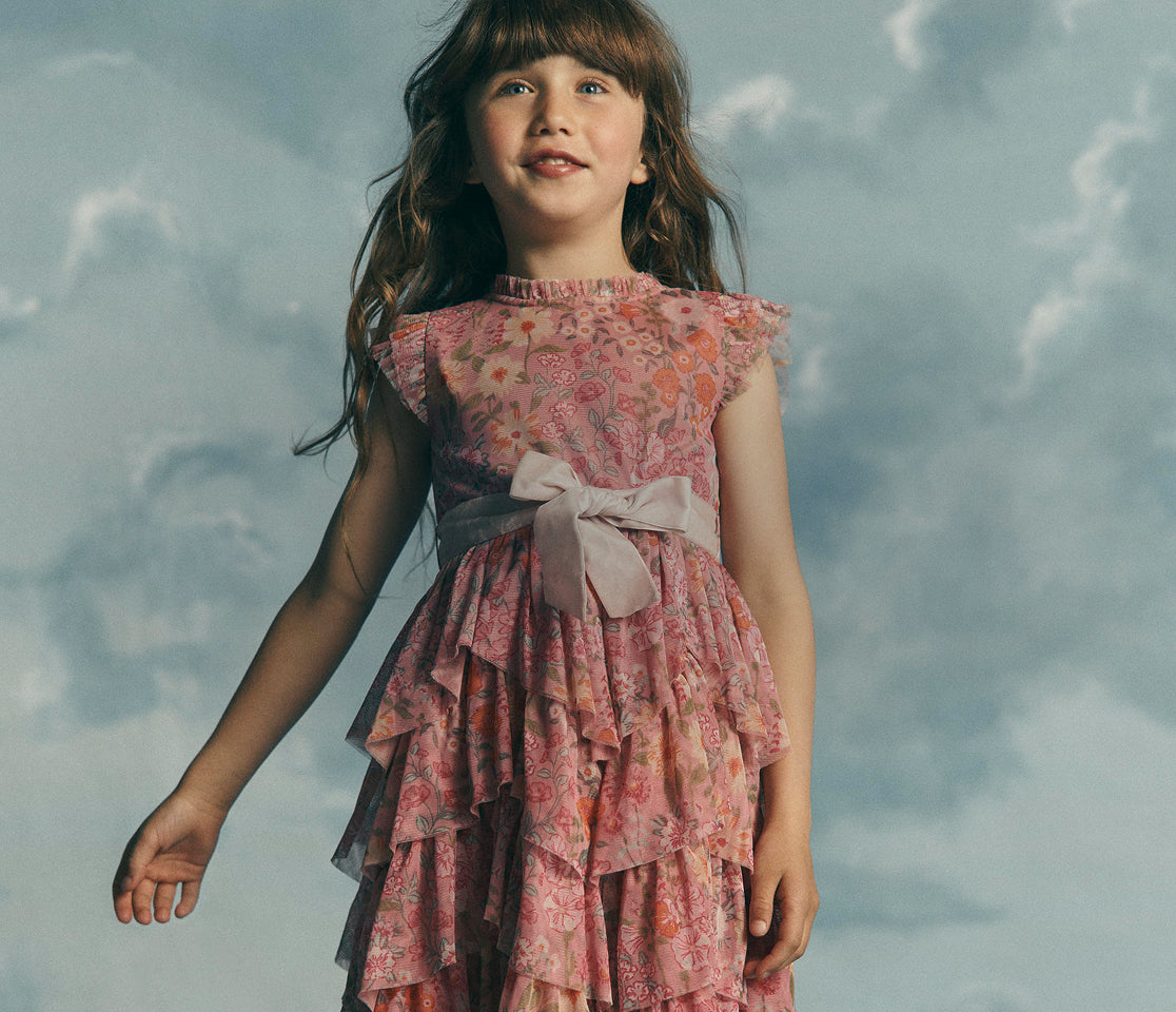 A young girl with long brown hair wearing a pink floral tiered dress with a large bow at the waist, standing against a cloudy sky background.