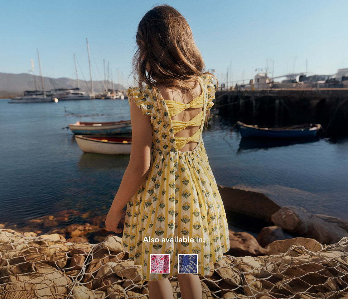 Young girl in a yellow dress standing by a harbor with boats and a clear sky.