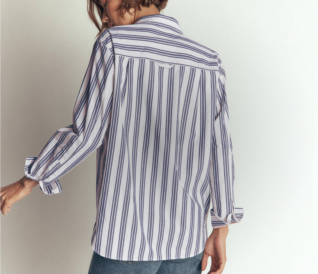 A woman wearing a white long-sleeve button-up shirt with vertical navy blue stripes, shown from a back-three-quarter view against a plain light gray background.