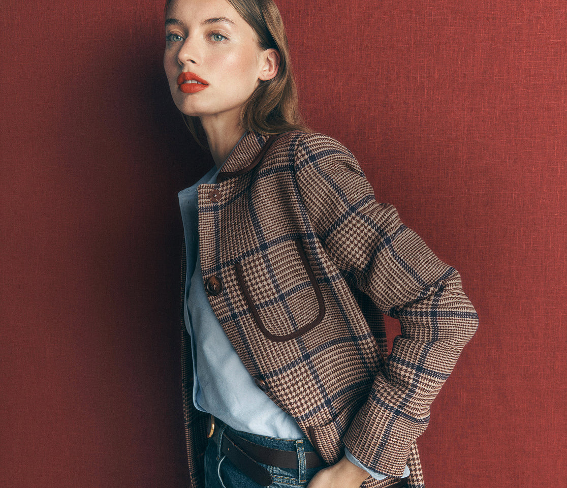 A woman wearing a beige and navy plaid blazer over a light blue shirt and dark wash jeans, posing against a textured dark red wall.