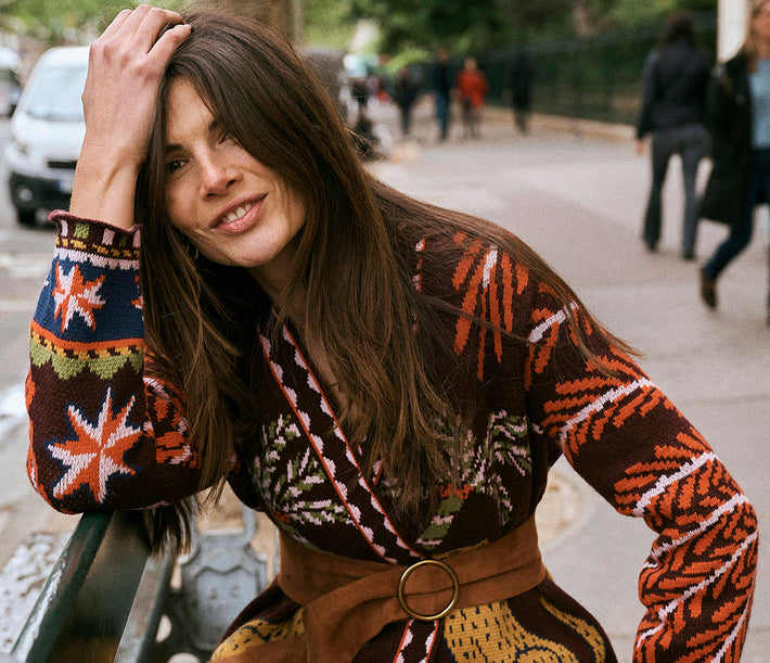 A woman outdoors wearing a brown knit wrap-style sweater with orange, cream, and navy geometric patterns, a wide brown belt, and a patterned skirt. She is leaning on a bench in a city setting with blurred pedestrians in the background.
