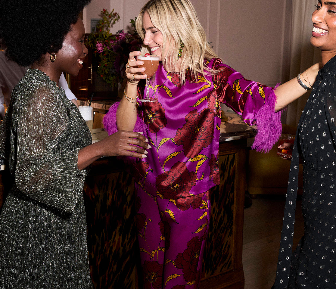 Three women socializing at a party, one in a purple outfit holding a drink.
