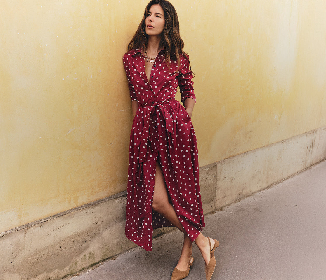 A woman wearing a long burgundy red and white polka dot midi dress with a tie waist, standing against a textured yellow wall.