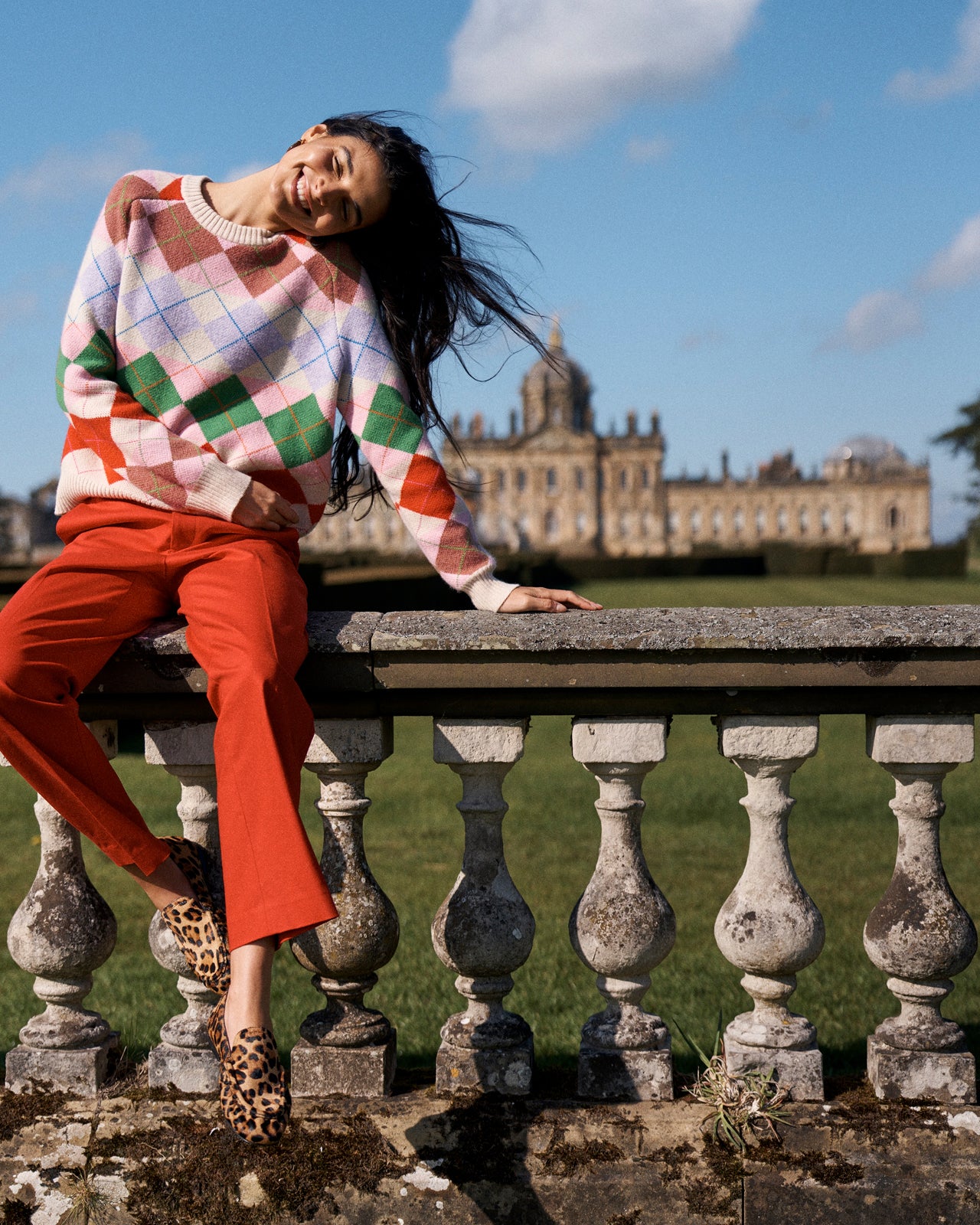 A woman wearing a multicolor argyle sweater, rust-orange cropped trousers, and leopard-print heels sits on a stone balustrade with a grand historic building and green lawn in the background.