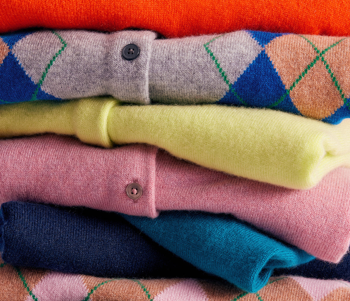 A close-up stack of folded knit cardigans in multiple colors, including orange, gray with argyle blue and peach, pale yellow, pink, navy, and teal.