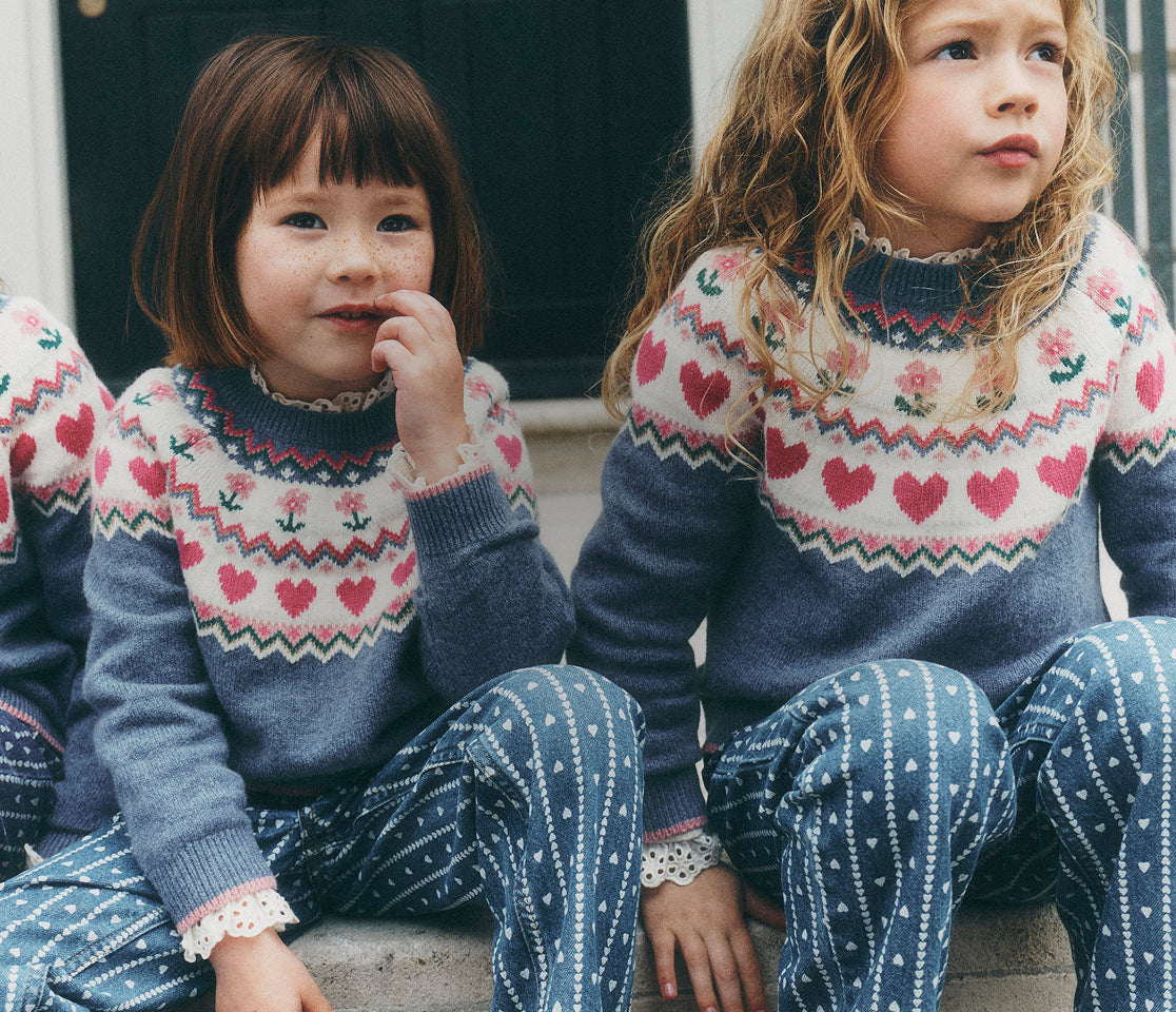 Two young girls sitting side by side wearing matching knit sweaters and patterned pants. The sweaters are navy blue with a cream yoke featuring pink hearts and red zigzag patterns, paired with navy pants that have white polka dots.