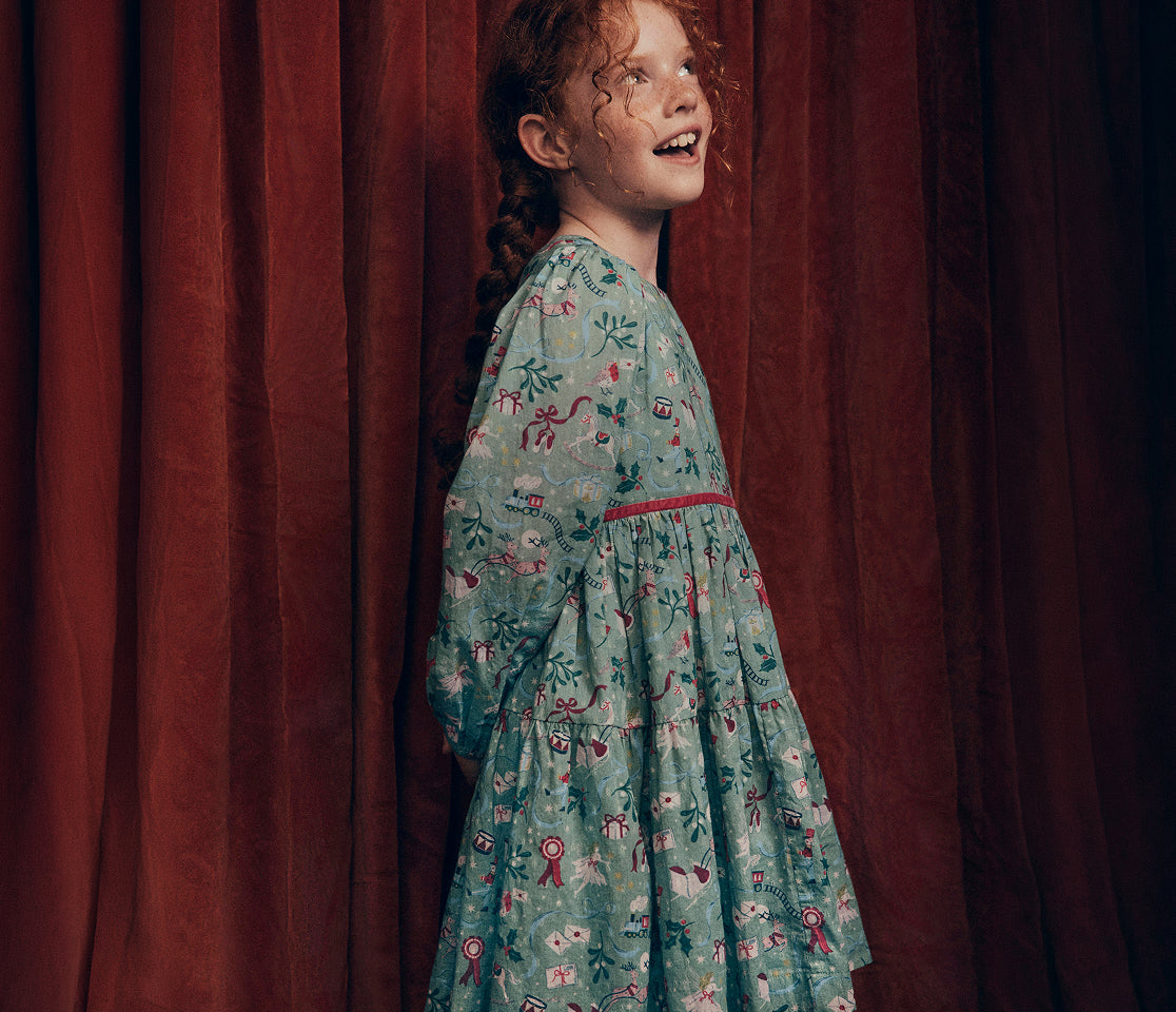 Child wearing a green long-sleeve dress with a festive illustrated print, gathered waist and tiered skirt