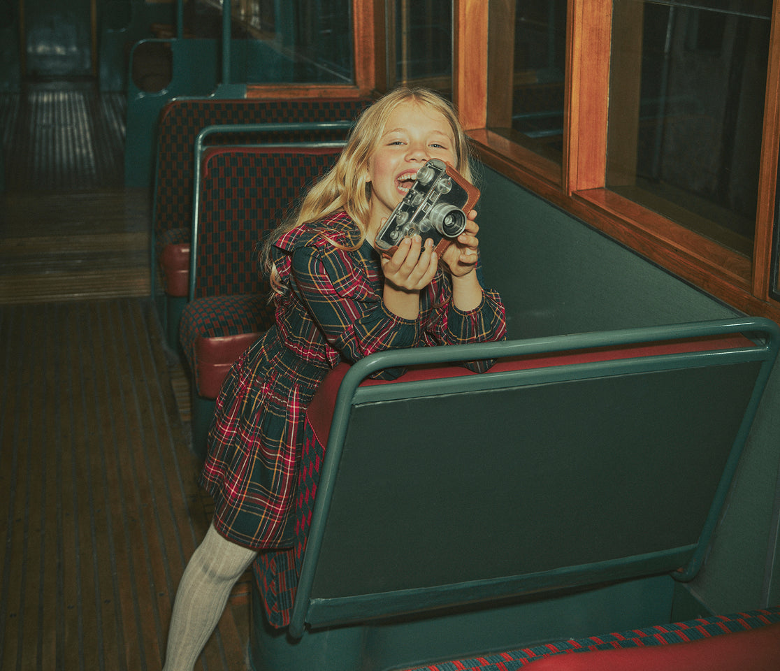 A young blonde girl in a red and black plaid dress sits on a vintage green bus seat, holding a retro silver camera to her face in a playful, candid pose.