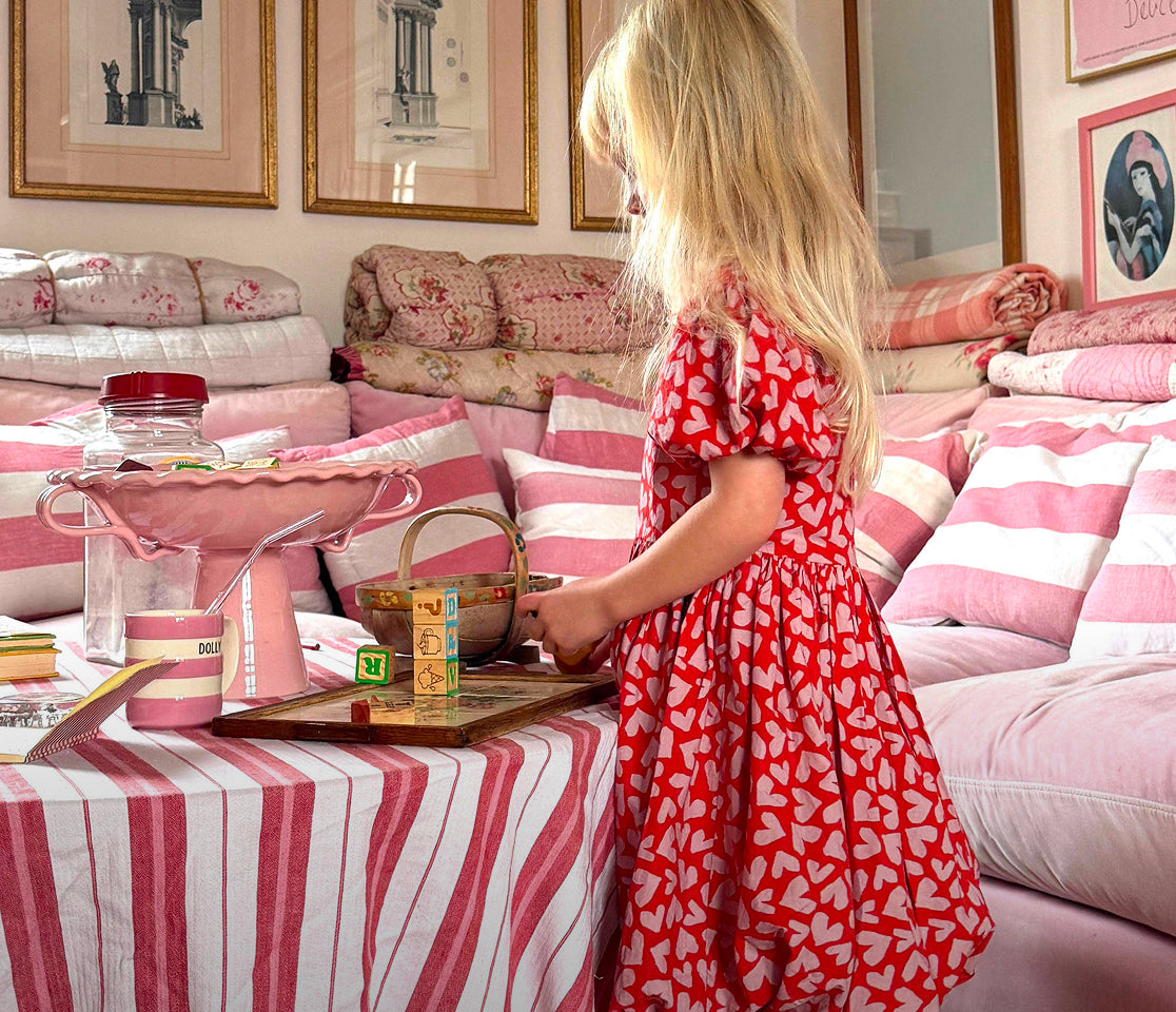Young girl in a red dress with white patterns standing in a room with pink and white striped furniture.