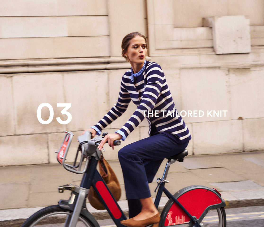 Woman riding a bicycle with a red basket in an urban setting, wearing a striped sweater.