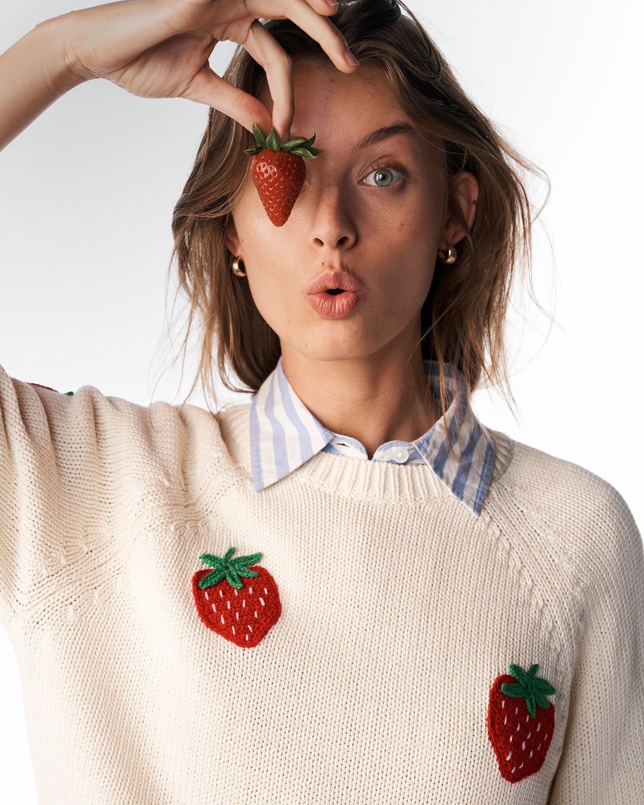 Woman wearing a cream sweater with strawberry designs, holding a strawberry to her eye.