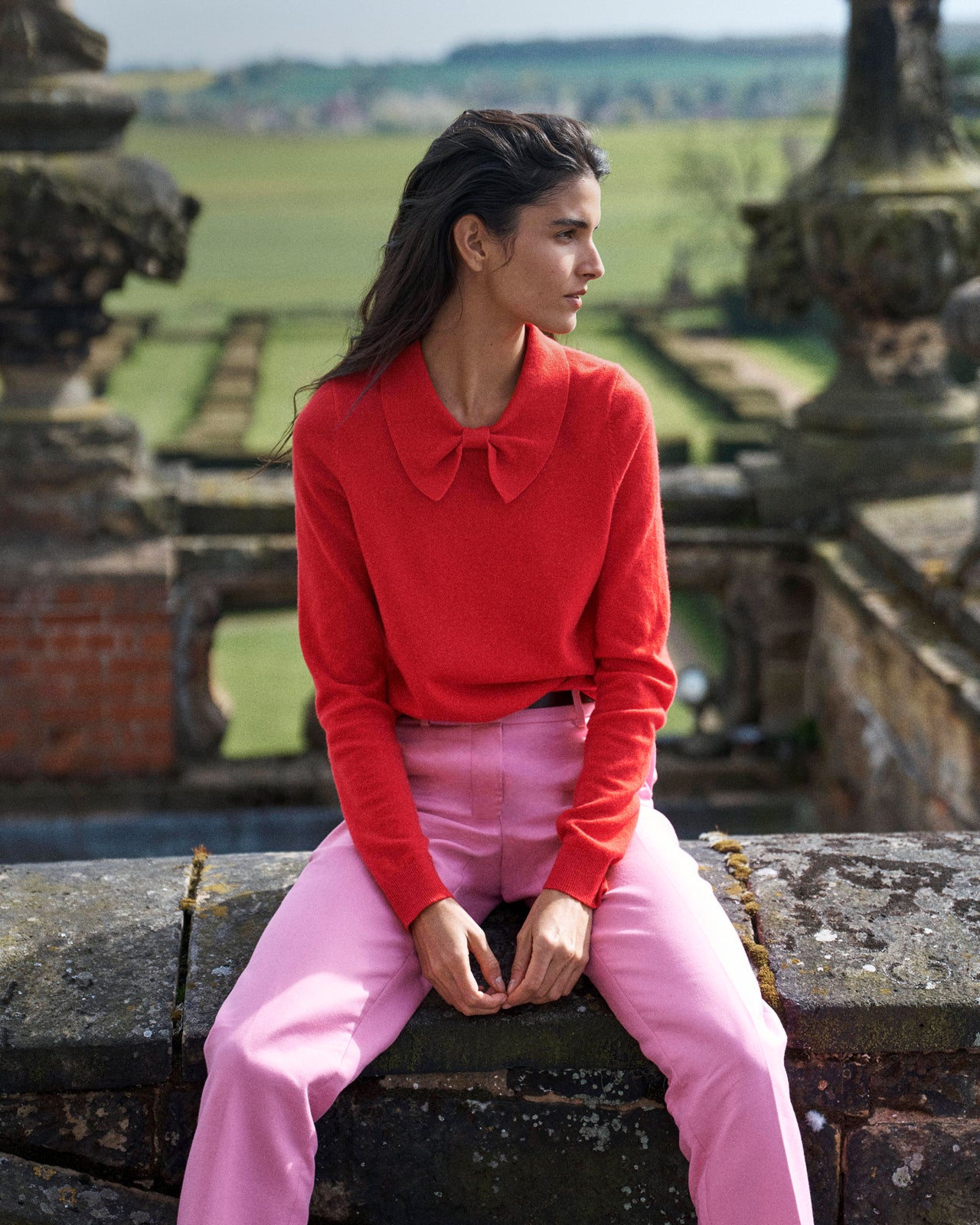 A woman sits on a stone wall in an outdoor garden setting, wearing a bright red long-sleeve knit top with a large bow collar and light pink trousers.