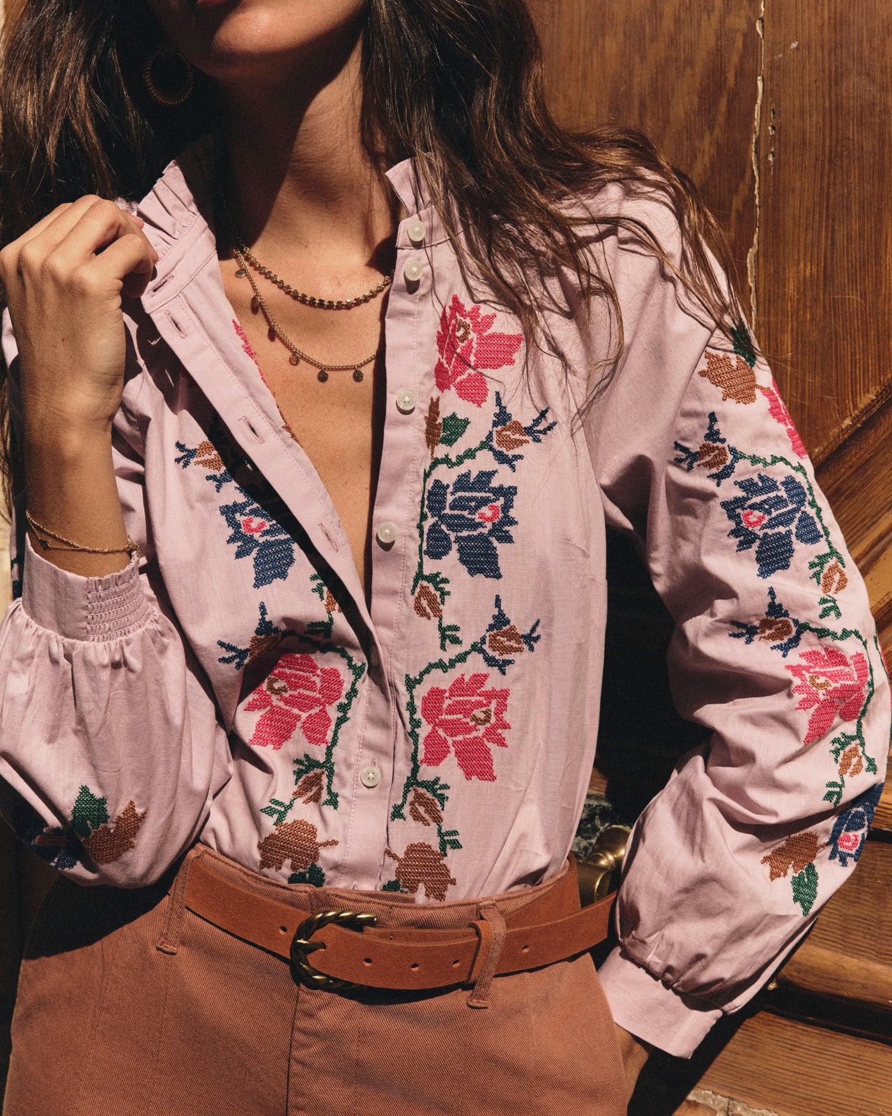 A woman wearing a light pink button-up blouse with colorful floral embroidery in pink, blue, green, and brown, paired with a brown belt and brown pants, set against a rustic wooden background.