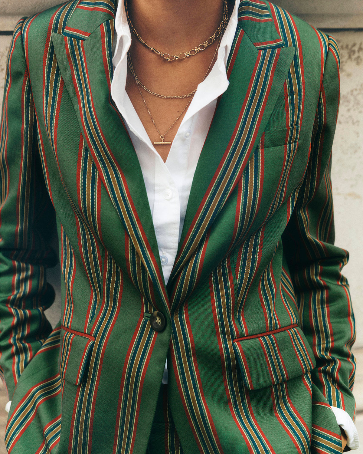 A close-up of a person wearing a green blazer with vertical red and blue stripes, layered over a white button-down shirt and gold necklaces.