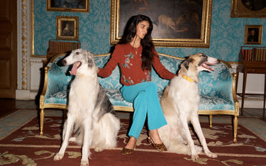 A woman sits on an ornate blue sofa flanked by two large grey and white dogs in a decorated room with blue patterned wallpaper and gold-framed paintings.
