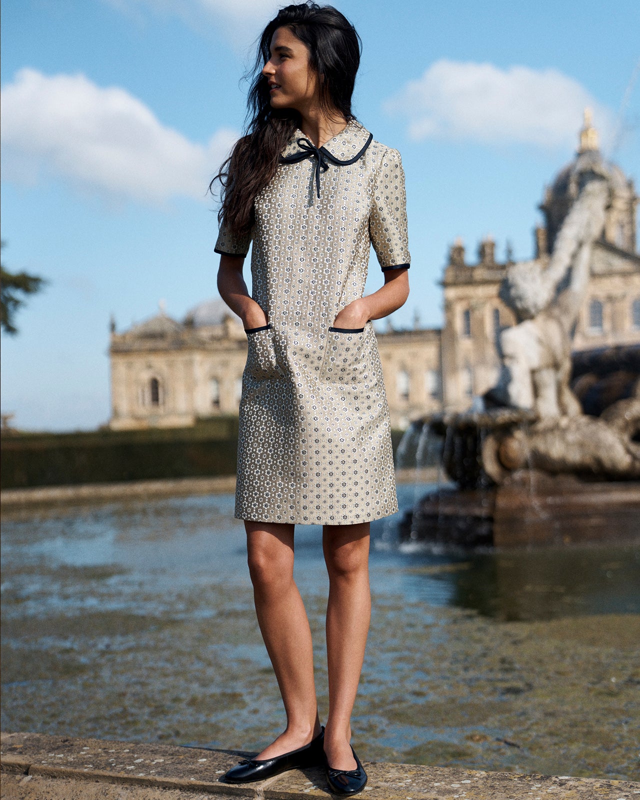 A woman stands outdoors in front of a fountain and grand building, wearing a short-sleeve beige dress with a black polka dot pattern, black collar with bow, and black flats.