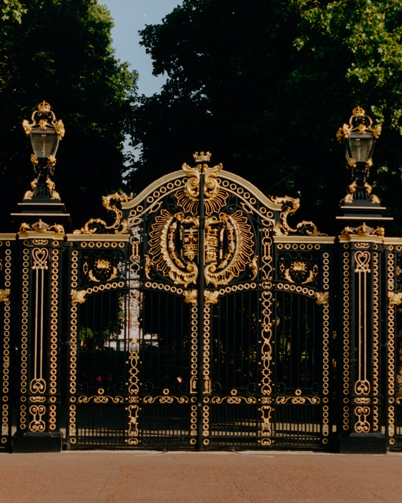 An ornate black wrought-iron gate with extensive gold decorative detailing, flanked by two lamp posts, set against a background of green trees and blue sky.