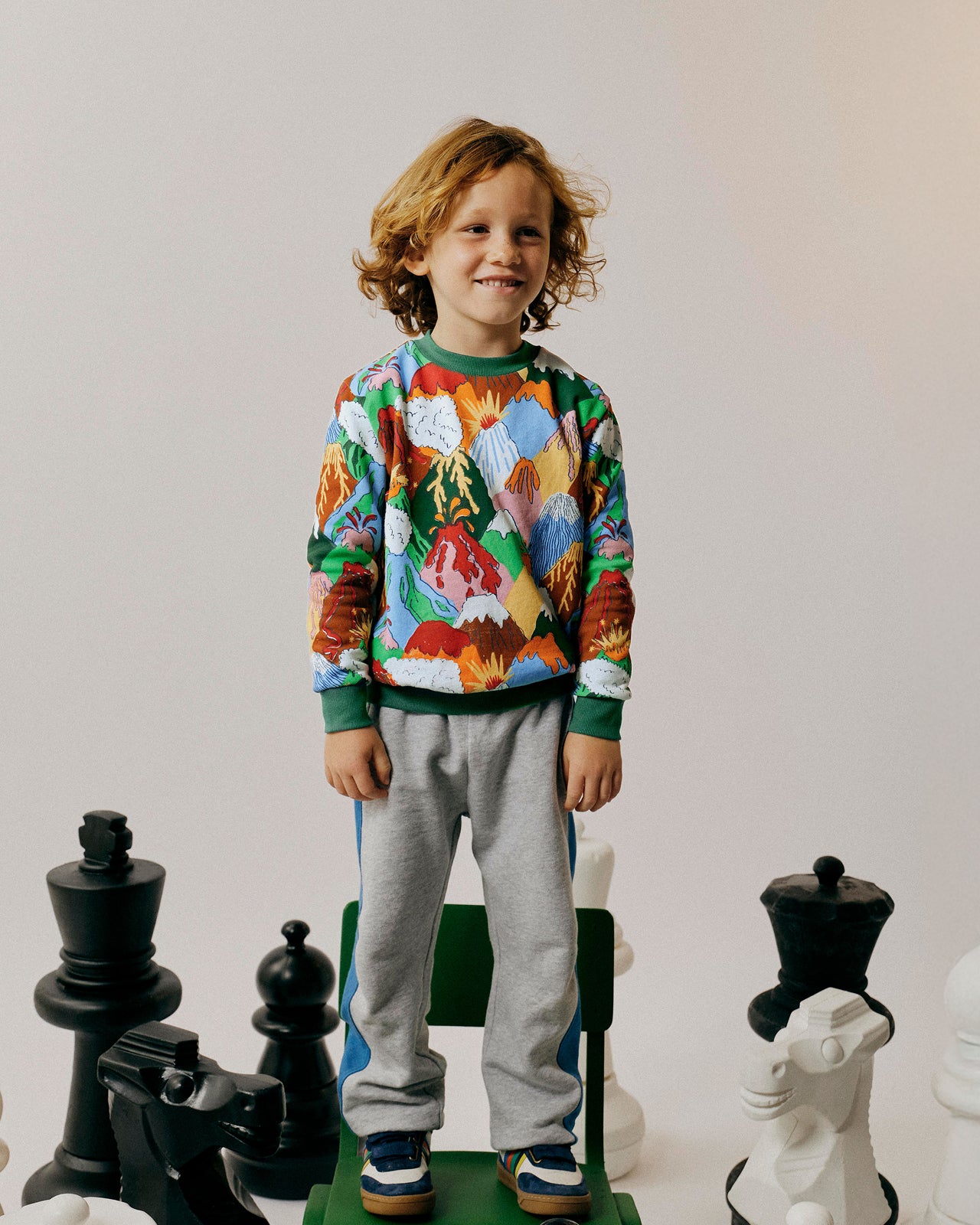 Child wearing a colorful sweater and gray pants standing on a green chair with chess pieces in the background.