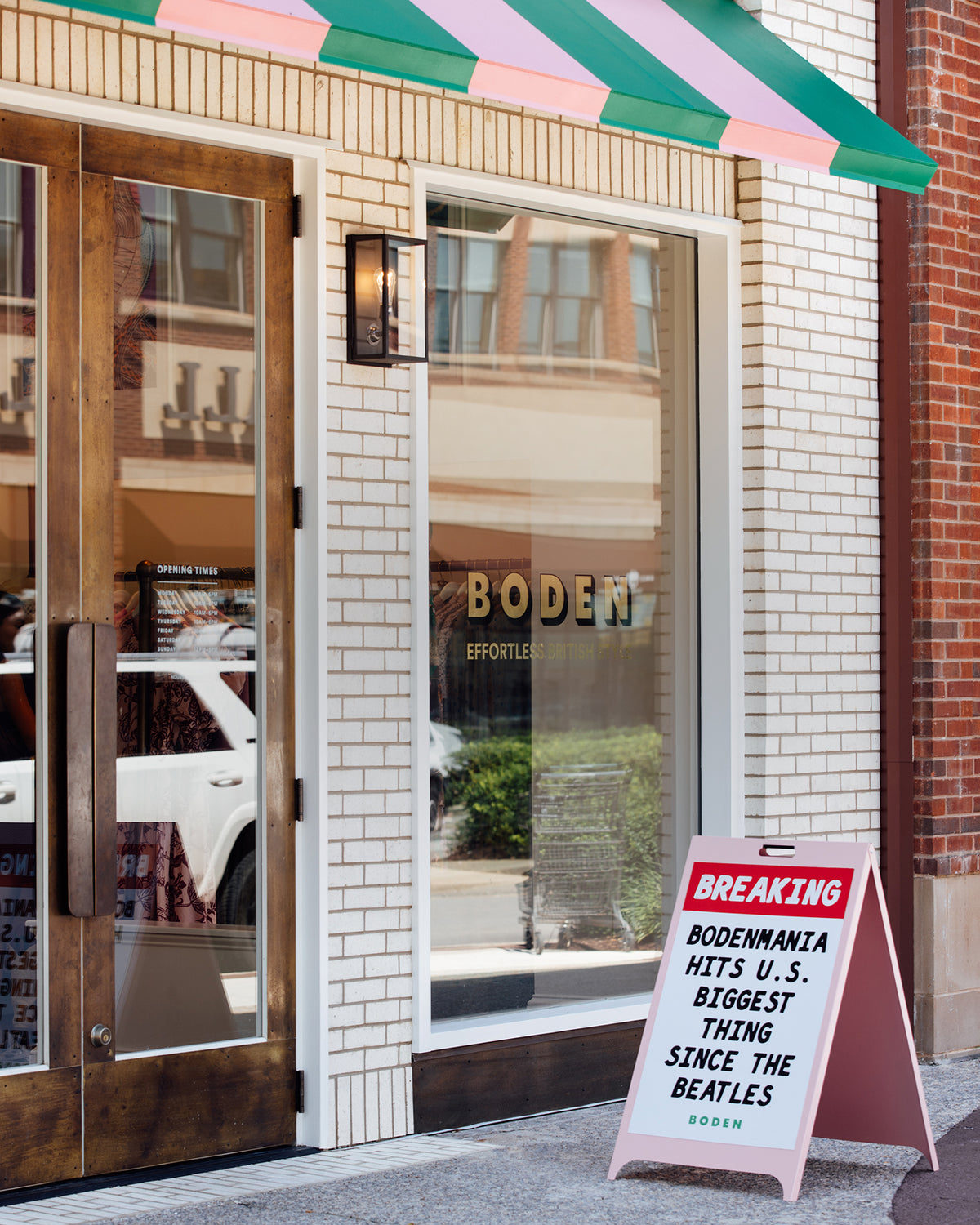 Boden Emporio storefront with a green and pink striped awning, white brick facade, glass doors, and a pink A-frame sidewalk sign reading 'BREAKING BODENWANTA HITS U.S.