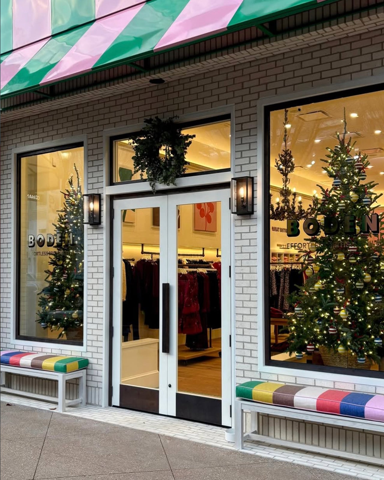 A storefront facade with white brick, a green and pink striped awning, double glass doors, large windows, bench seating with colorful striped cushions, and Christmas trees visible inside.