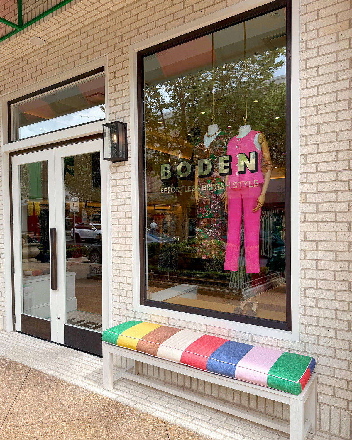 A storefront window display for Boden, featuring a bright pink outfit on a mannequin, with a colorful striped bench in front of a beige brick building.