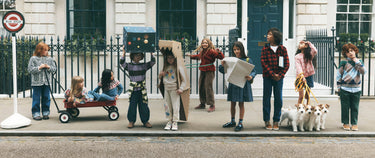 A group of children standing on a street sidewalk in front of a building, many wearing creative costumes like cardboard boxes and holding a red wagon, with two small dogs beside them.