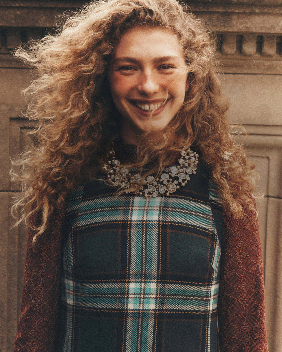 Person with long, curly hair standing in front of a stone wall, wearing a dark green and blue plaid sleeveless dress layered over a burgundy lace top, accessorized with a large jeweled statement necklace. The portrait is warmly lit, with hair blowing slightly across the face.