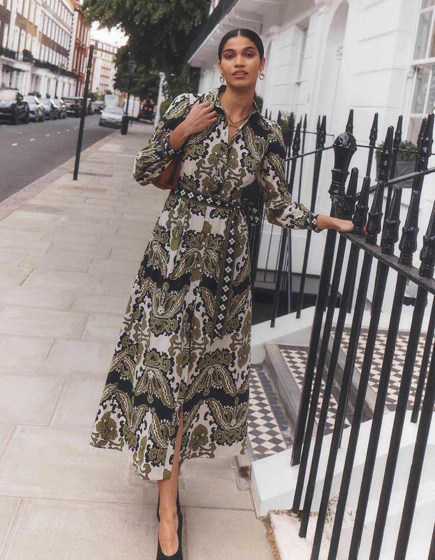 A woman models a long-sleeved midi dress with a green, black, and white paisley pattern, standing on a city sidewalk next to a black metal fence.
