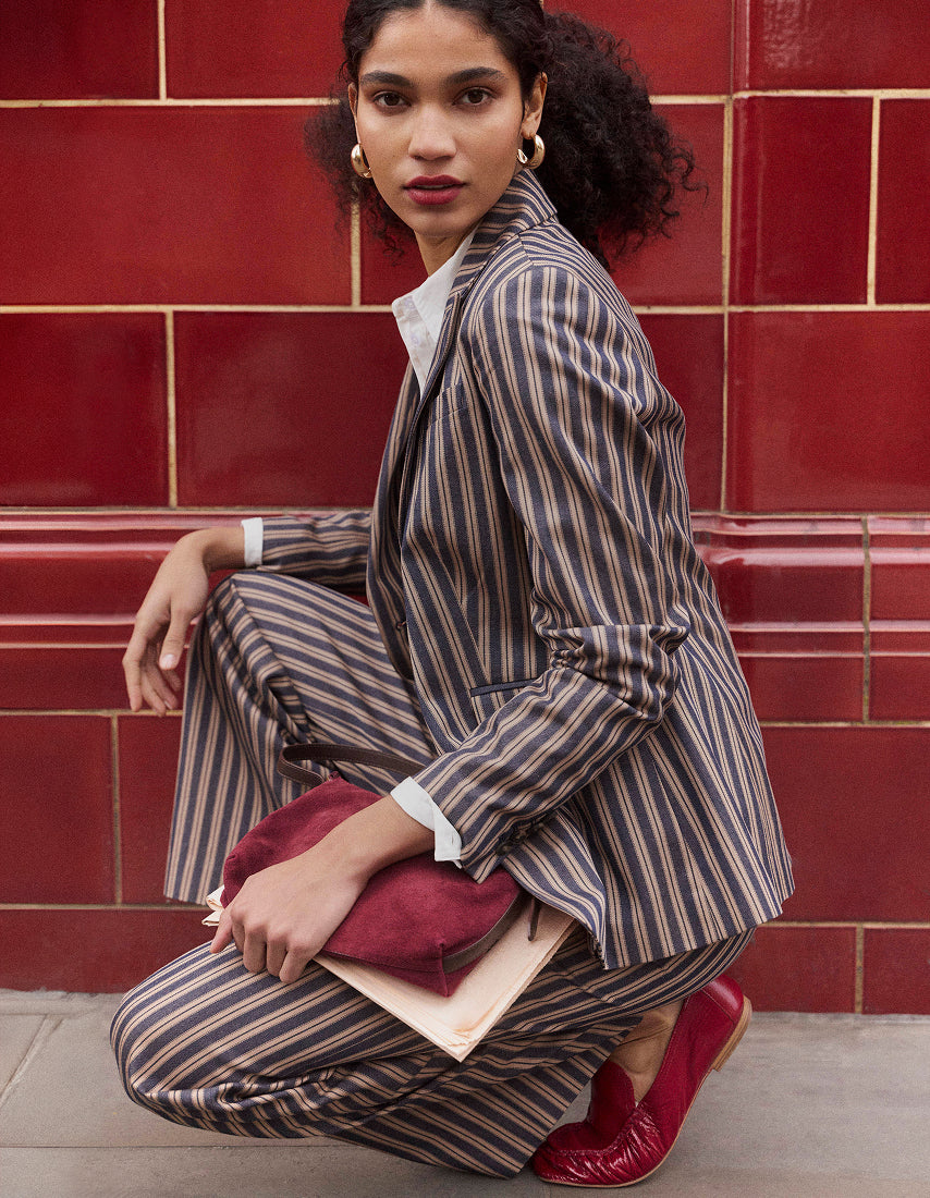 A model crouches in front of a red tiled wall wearing a brown and beige pinstripe blazer and matching trousers, holding a burgundy clutch and a light-colored book, with red leather loafers.