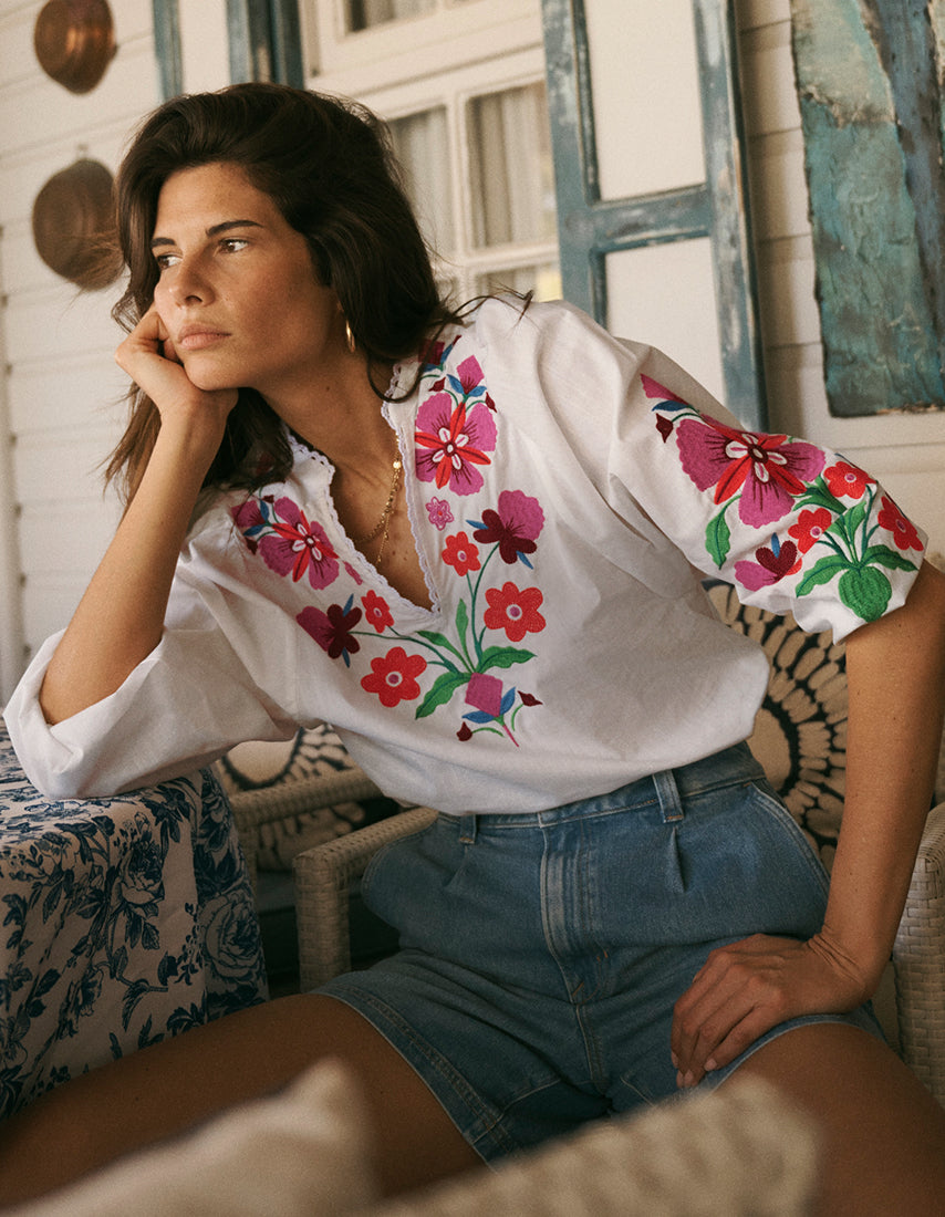 Woman wearing a white blouse with colorful floral embroidery and denim shorts, sitting in a decorative room.