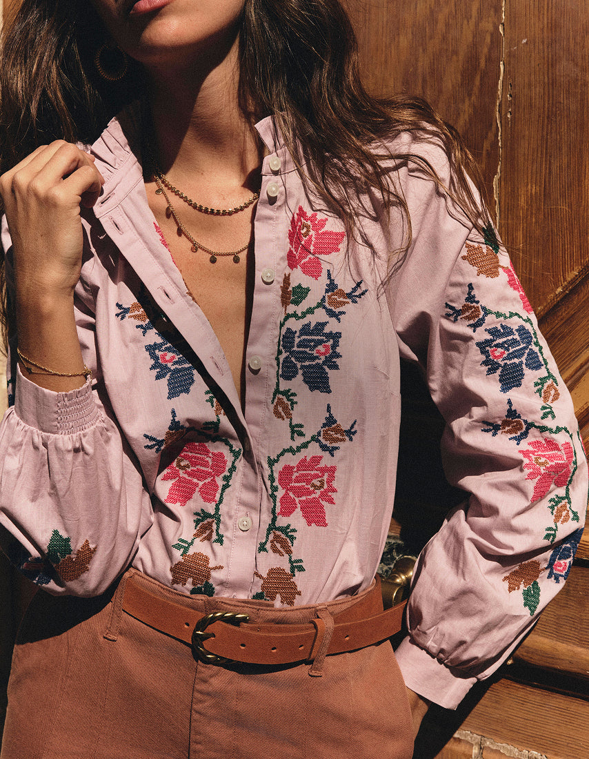 A woman wearing a light pink button-up blouse with colorful floral embroidery, paired with a brown belt and brown pants, posed against a rustic wooden background.