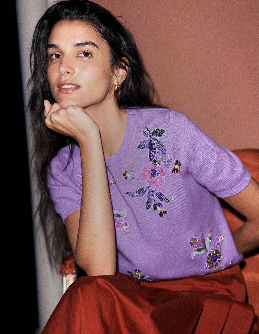 A woman posing in a lavender short-sleeve knit sweater with colorful floral embroidery and a rust-colored satin skirt, set against a warm terracotta background.