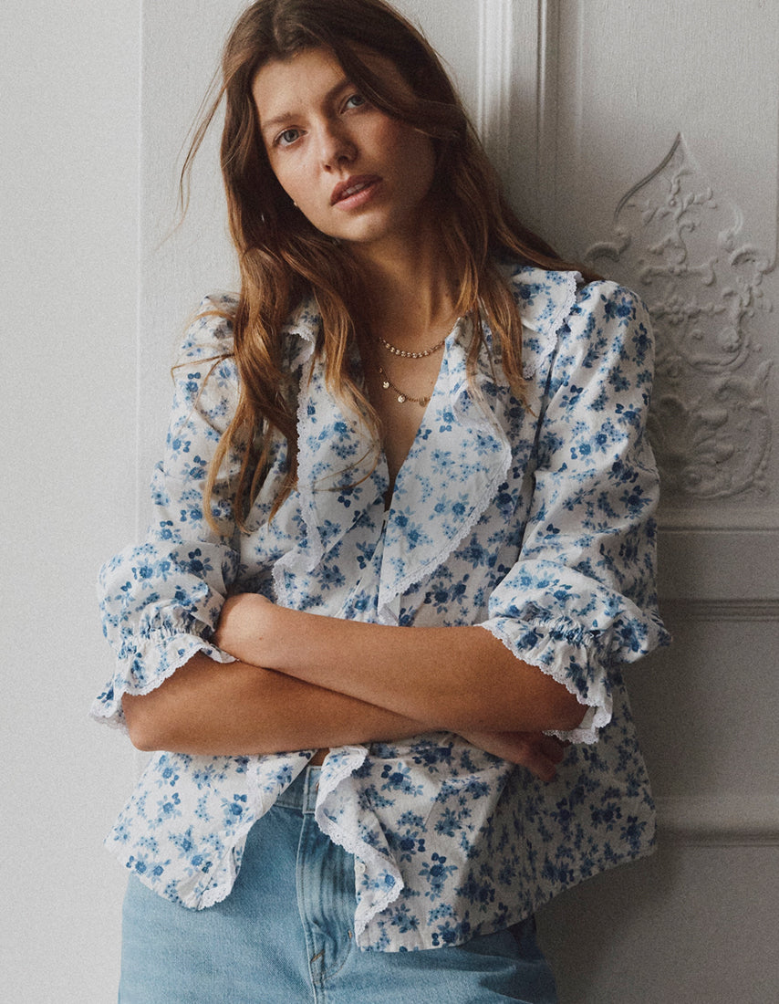 Woman wearing a blue floral blouse and jeans against a light-colored wall.