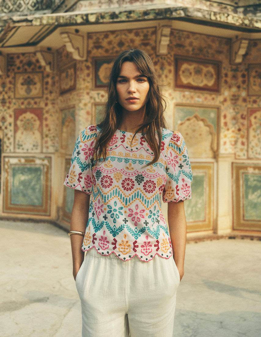 Woman wearing a colorful patterned top and white pants standing in front of an ornate wall.