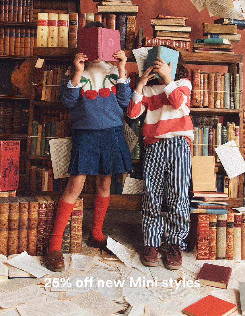 Two children standing in a book-filled room, each holding a book over their face. The child on the left wears a blue sweater with a cherry motif, a blue pleated skirt, and red knee-high socks.