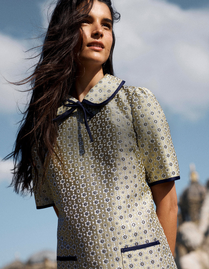 A woman wearing a short-sleeved cream-colored dress with a small blue and green floral pattern, a dark navy collar with a tie, and navy trim, photographed outdoors against a blue sky.
