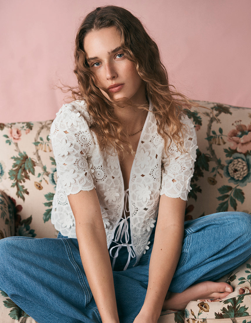 Woman wearing a white lace top and blue jeans sitting on a floral couch.