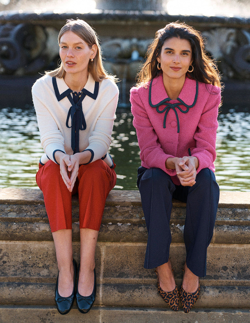 Two women sitting side-by-side on stone steps in front of a fountain. The woman on the left wears a cream long-sleeve blouse with a black bow collar, rust-red cropped trousers, and dark teal heels.