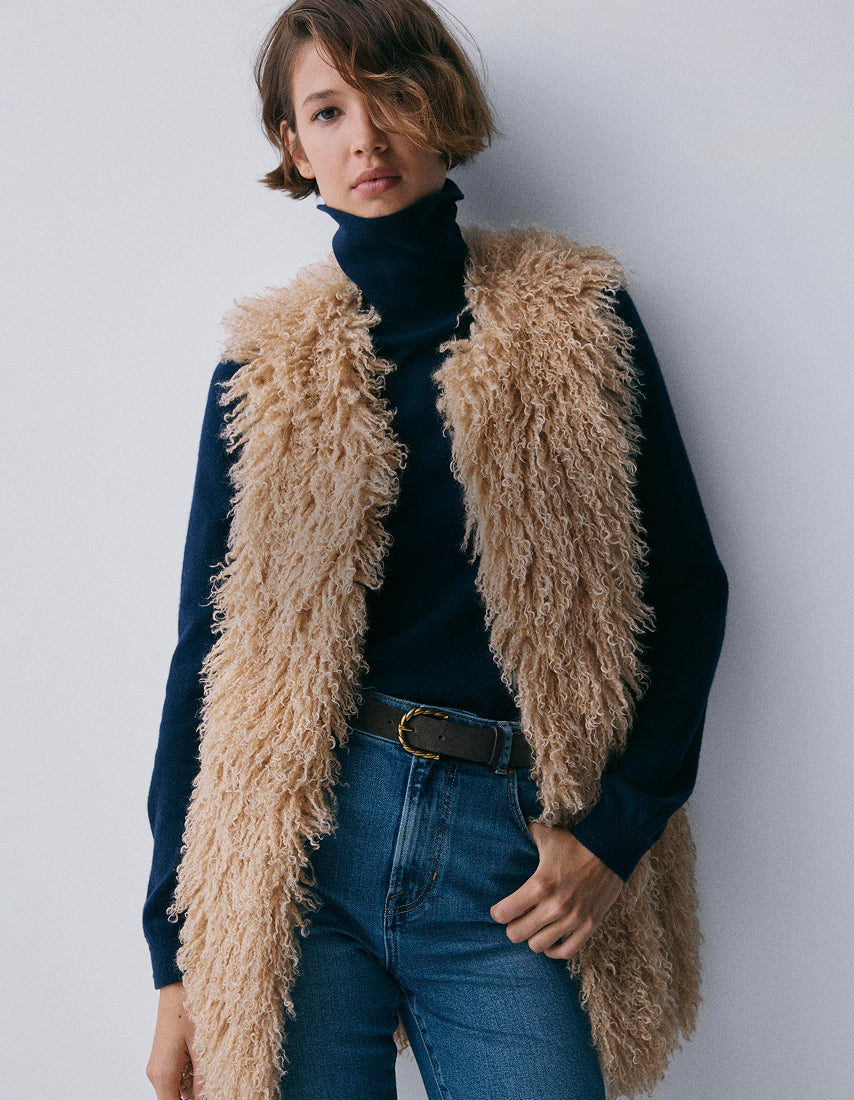 Person wearing a fluffy beige vest over a dark turtleneck and jeans against a plain background
