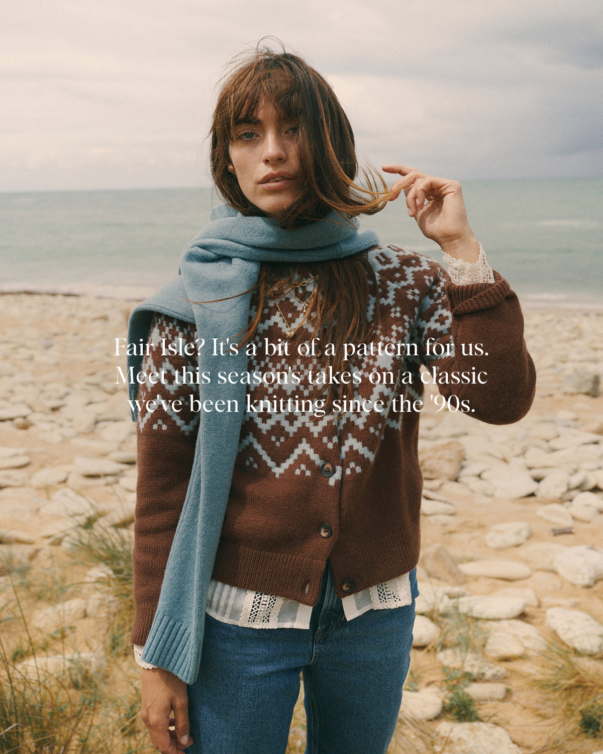 A woman stands on a rocky beach wearing a brown knit cardigan with a white geometric pattern, a light blue knit scarf, and blue jeans. White text across the center reads, 'Fair Isle? It's a bit of a pattern for us.