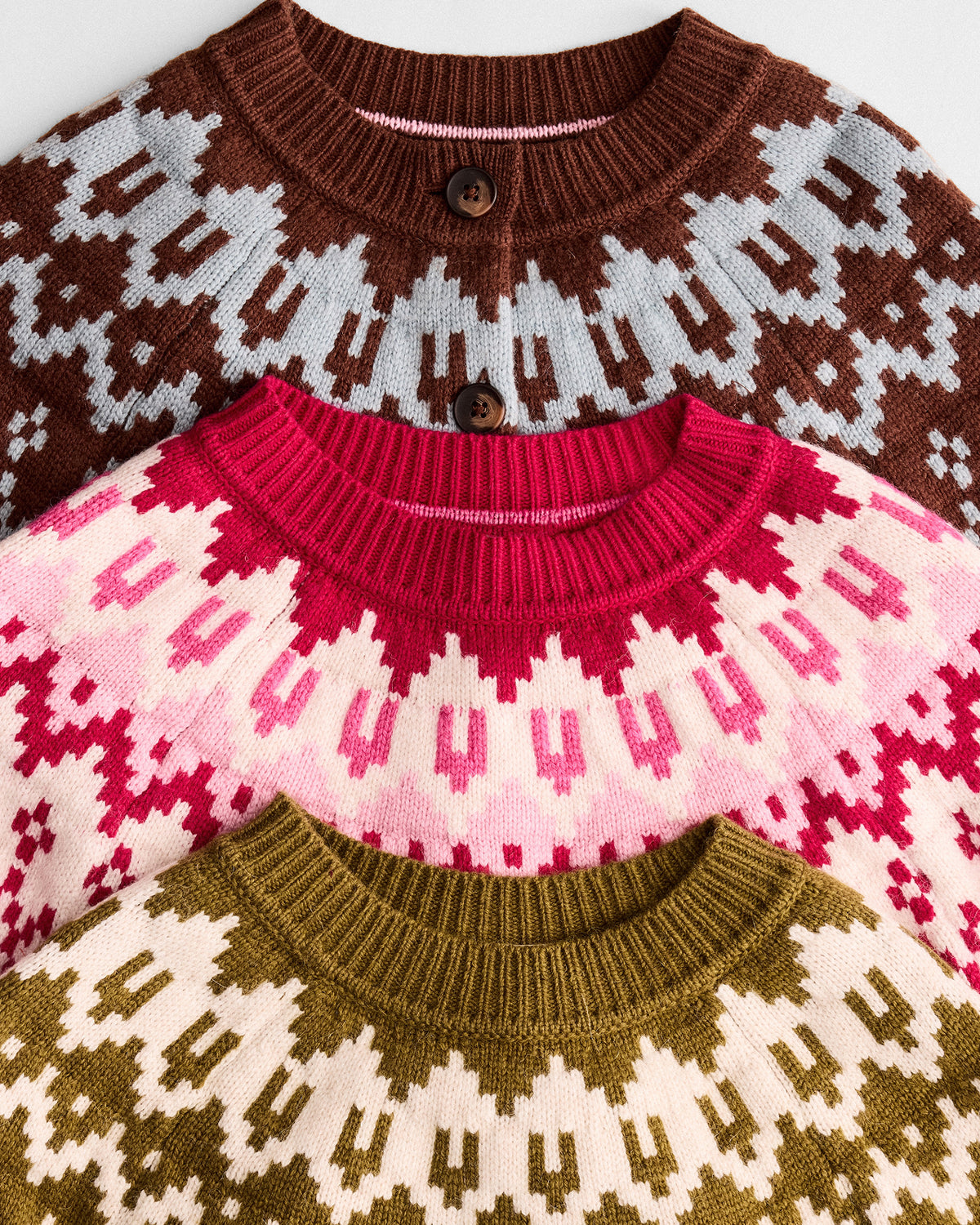 Three knit crewneck cardigans layered together, each with a geometric Fair Isle pattern: brown and light gray, bright pink and cream, and olive green and cream.