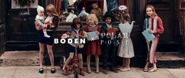 Group of children gathered outside a festive shop window decorated with holiday cards, gifts and toys. One child stands with a scooter, another holds a baby, while others carry cards and letters. The children wear colourful seasonal outfits, including a patterned dress, a checked suit, angel wings and a striped knit set. Visible text reads “MINI BODEN x POLAR POST.”